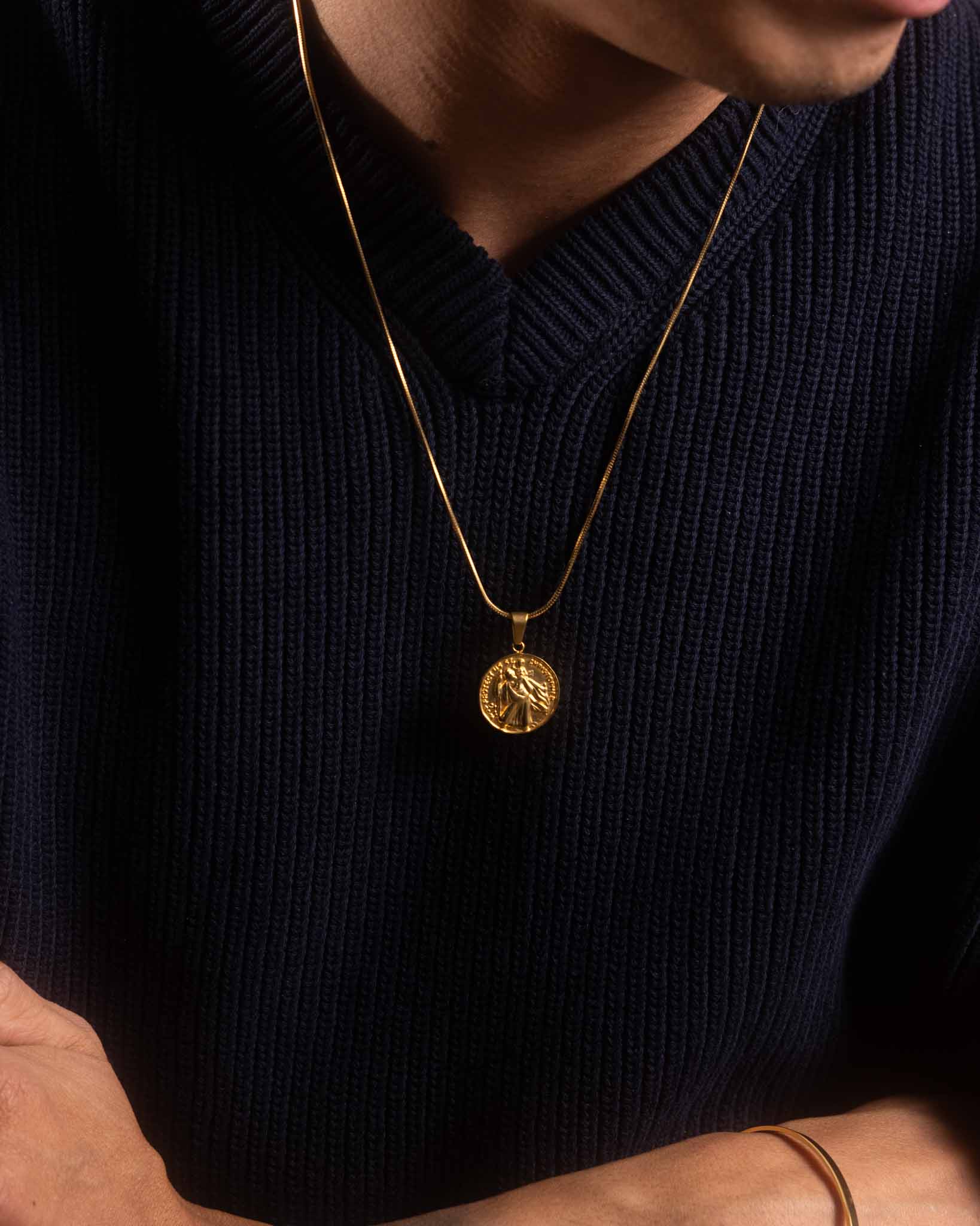 En person i mörk ribbstickad tröja korsar armarna och bär ett herrhalsband i guld med ett runt, upphöjt hänge.