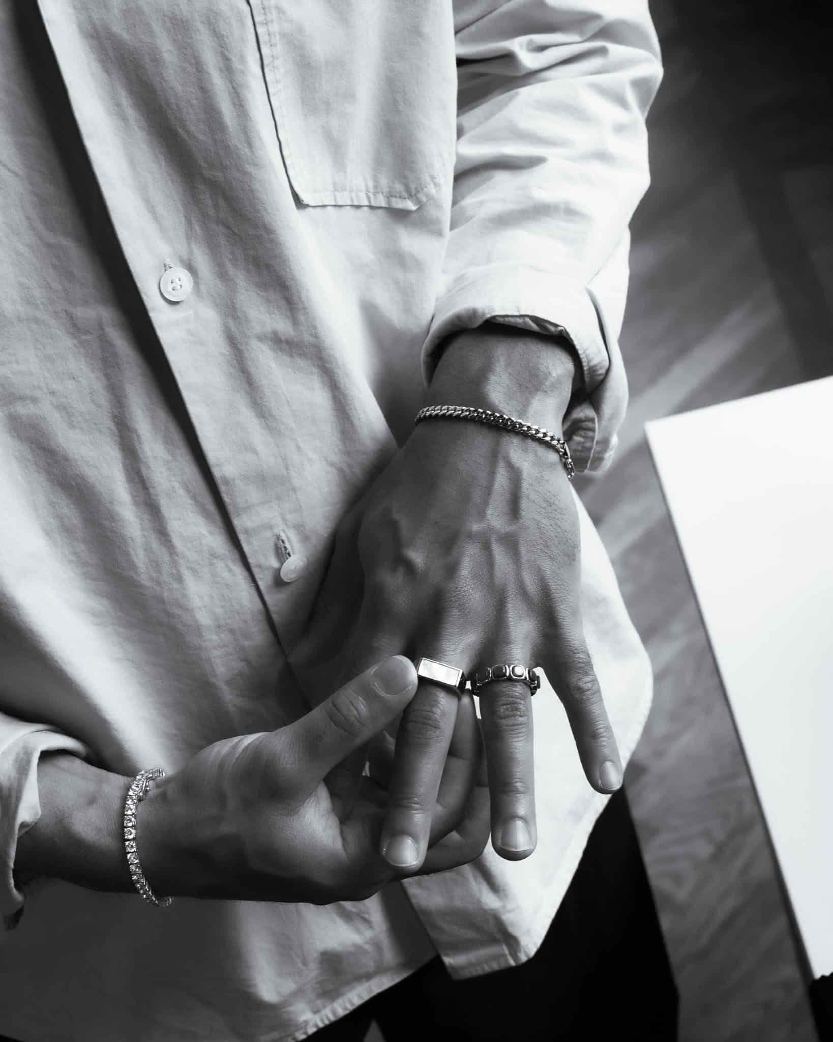 Svartvitt foto som visar en persons händer och eleganta smycken, med ett herrarmband i pansarlänkdesign tillsammans med en knäppt skjorta, armband och ringar när en hand justerar en ring på den andra.