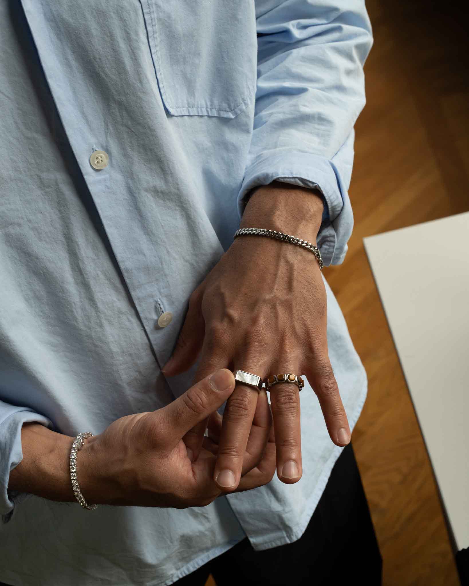 En person i en ljusblå skjorta med knappar visar sina händer dekorerade med silverringar och Pansar-armbandet i silver på 5 mm, med trägolv i bakgrunden.