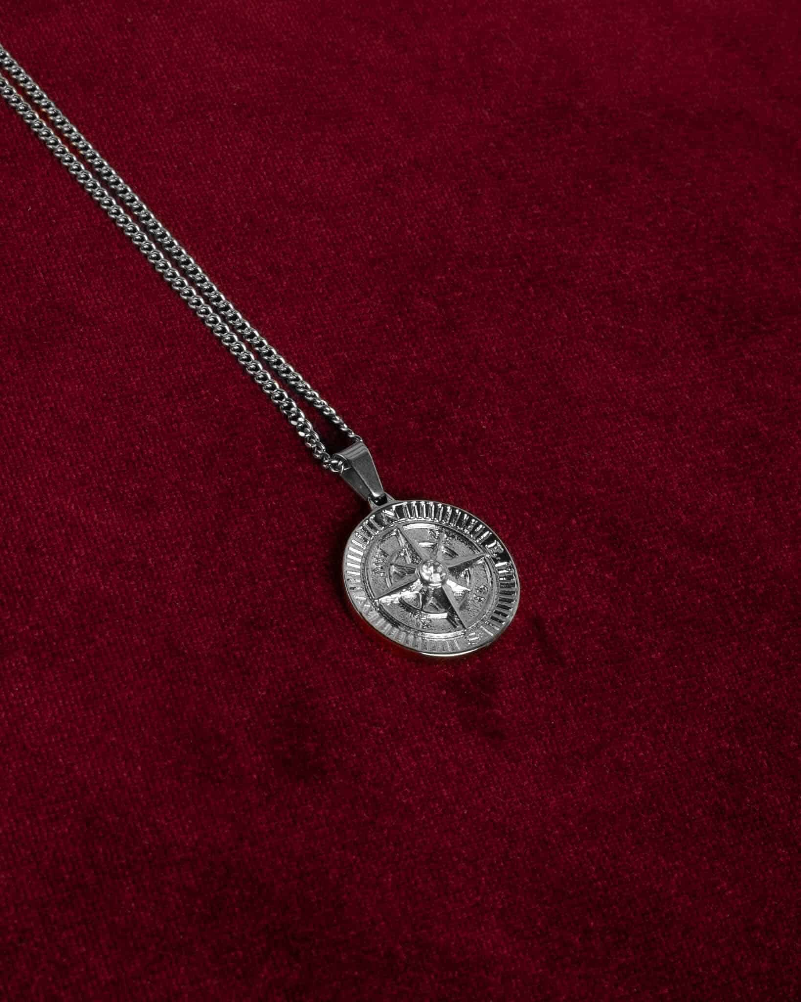 Ett Kompass halsband med silverhänge och en rund medaljong med intrikat design, placerat på en rik vinröd sammetsbakgrund.