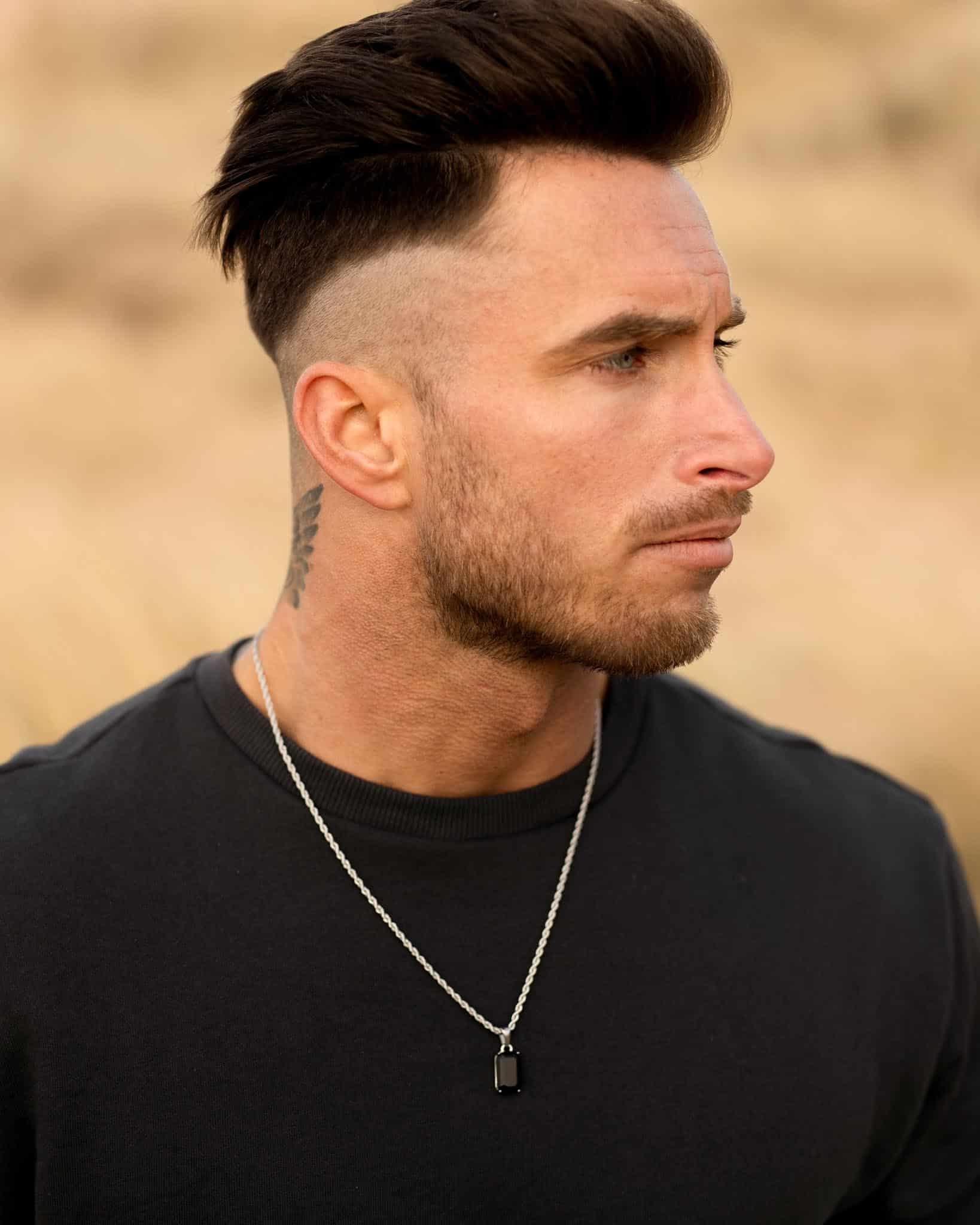 En man med välfriserat mörkt hår, vältrimmat skägg och en synlig tatuering i nacken bär en svart skjorta och ett svart onyxhalsband i silver och tittar åt sidan mot en suddig bakgrund utomhus.