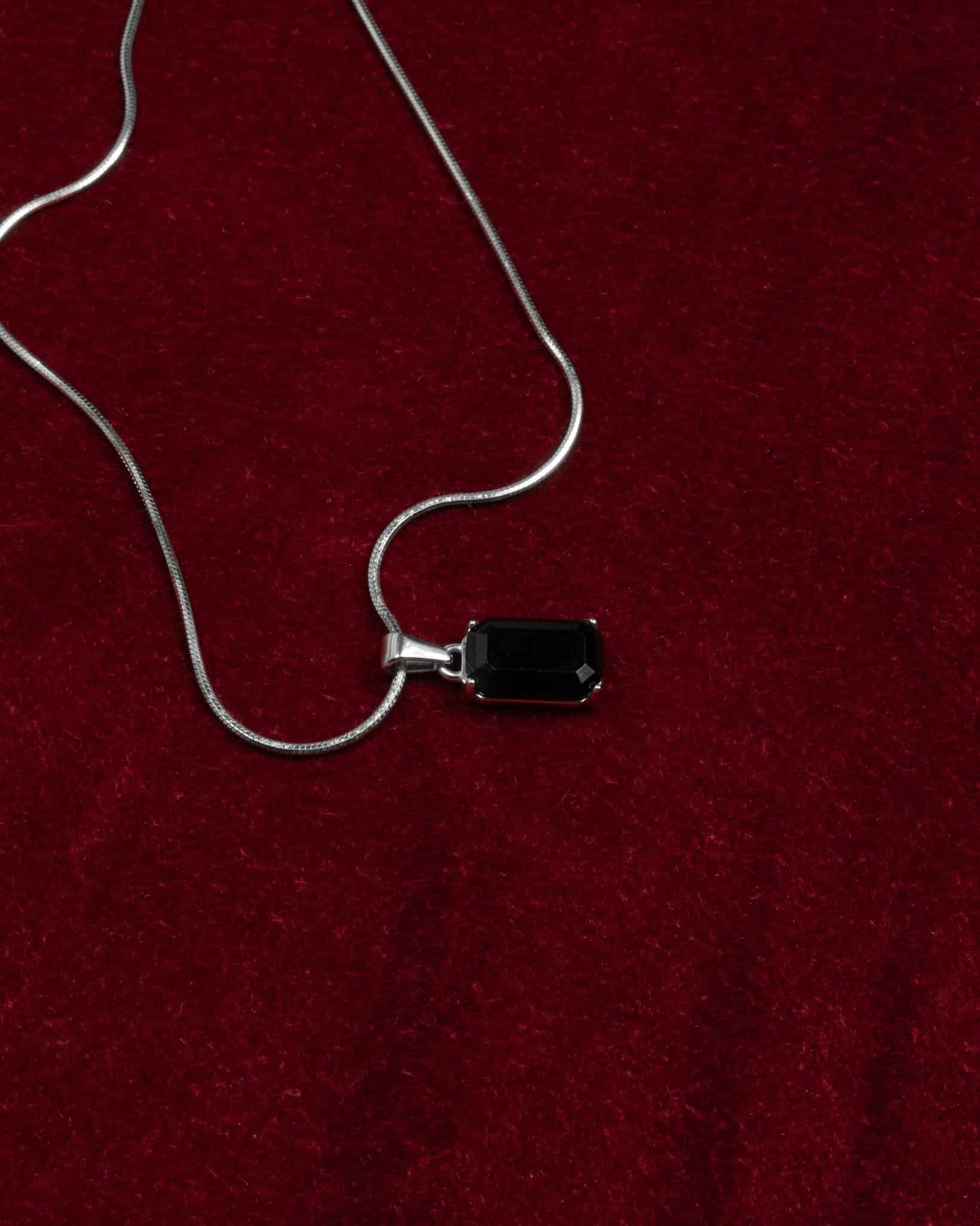 Ett silverhalsband i svart onyx med ett rektangulärt ädelstenshänge visas på en rik, mörkröd bakgrund av sammetstyg.