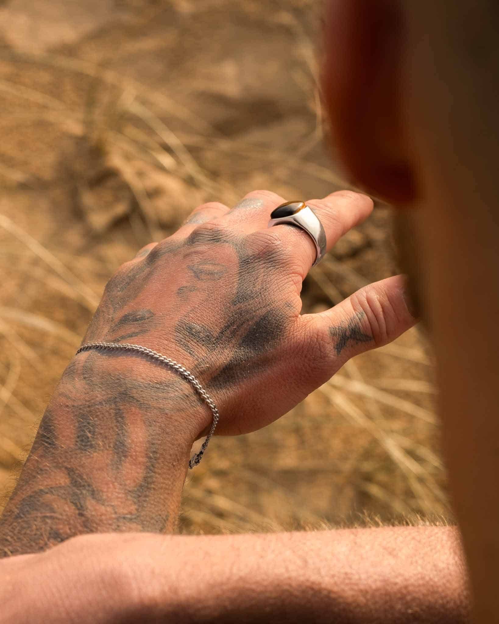 En tatuerad hand som bär en silverring och Pansararmbandet silver 3mm visas i närbild mot en suddig sandig utomhusbakgrund, med bärarens huvud och axel synliga i förgrunden.