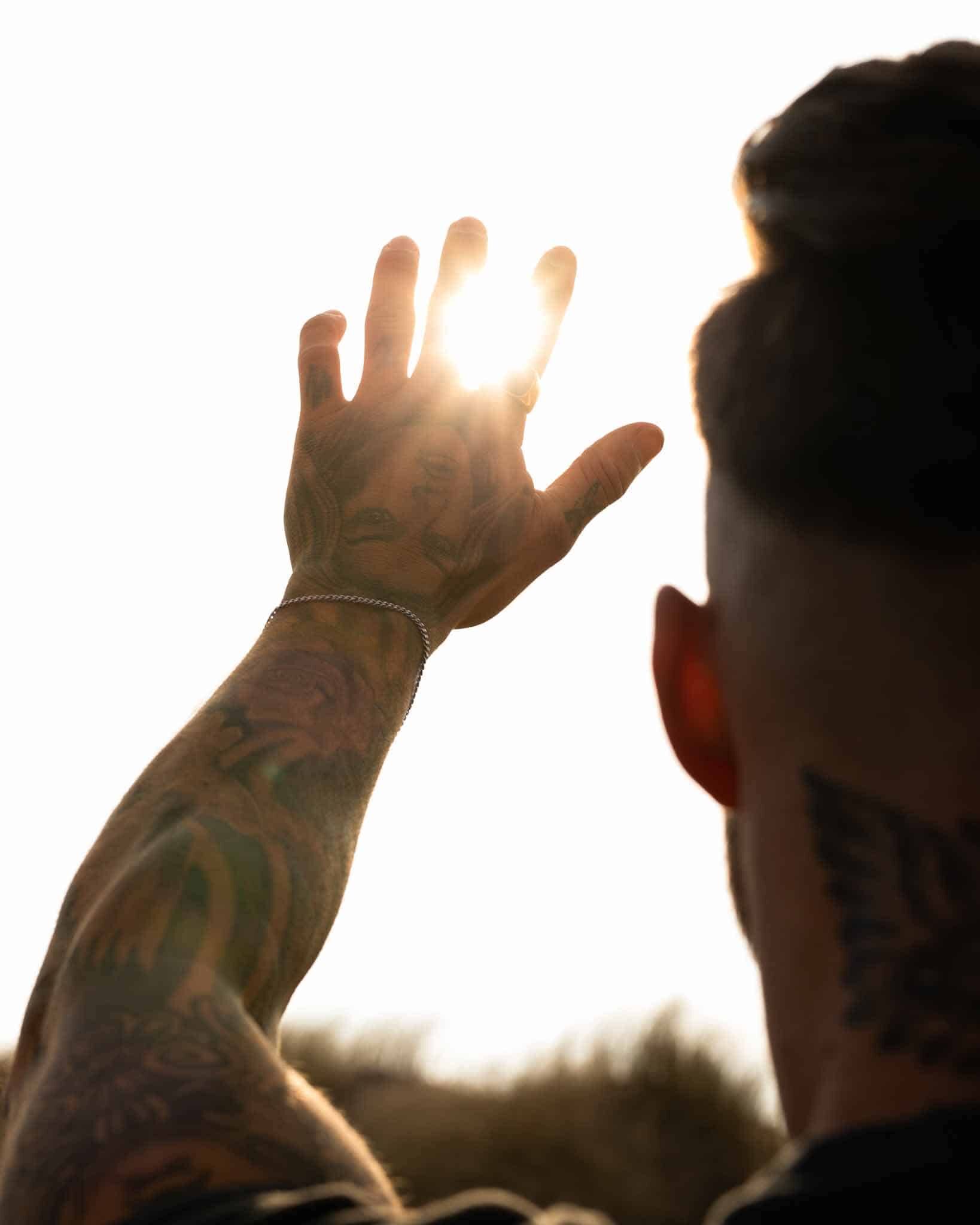 En tatuerad person som bär ett Pansararmband silver 3mm lyfter handen mot starkt solljus, fingrarna är utspridda när strålarna passerar mellan dem. I den bakgrundsbelysta scenen syns endast armen och sidan av huvudet i silhuett.