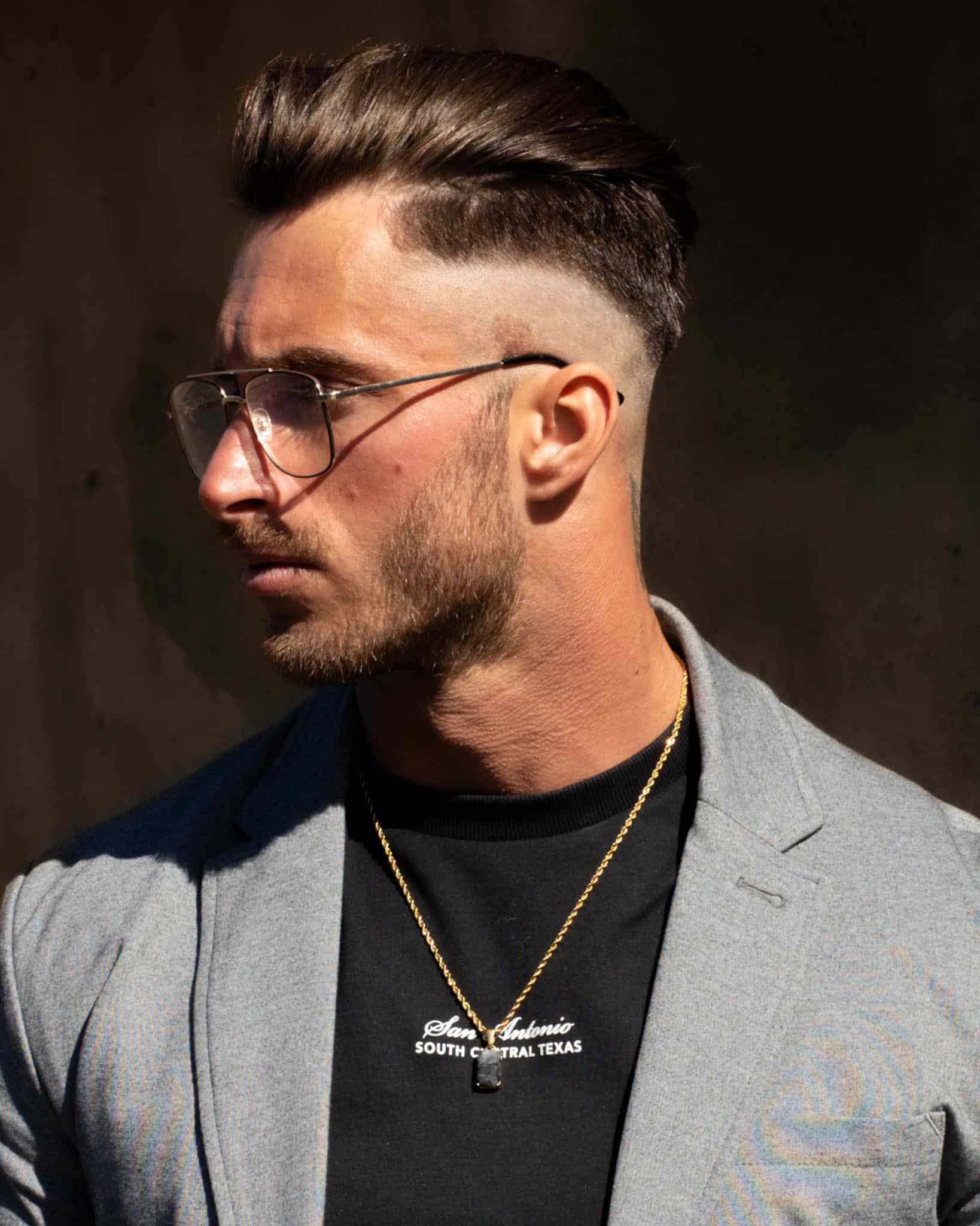 En man med stylat brunt hår, skägg och glasögon bär en grå kavaj, svart skjorta och halsbandet Gemstone Black Onyx Marbled Guld. Han står mot en mörk bakgrund och tittar åt vänster.