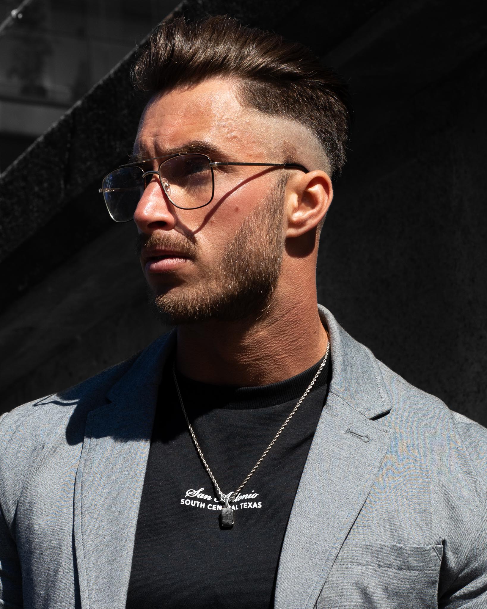 En man med kort hår, trimmat skägg och glasögon bär en ljusgrå kavaj, svart skjorta och halsbandet Gemstone Black Onyx Marbled Silver. Han står i solljus mot en mörk, suddig bakgrund.