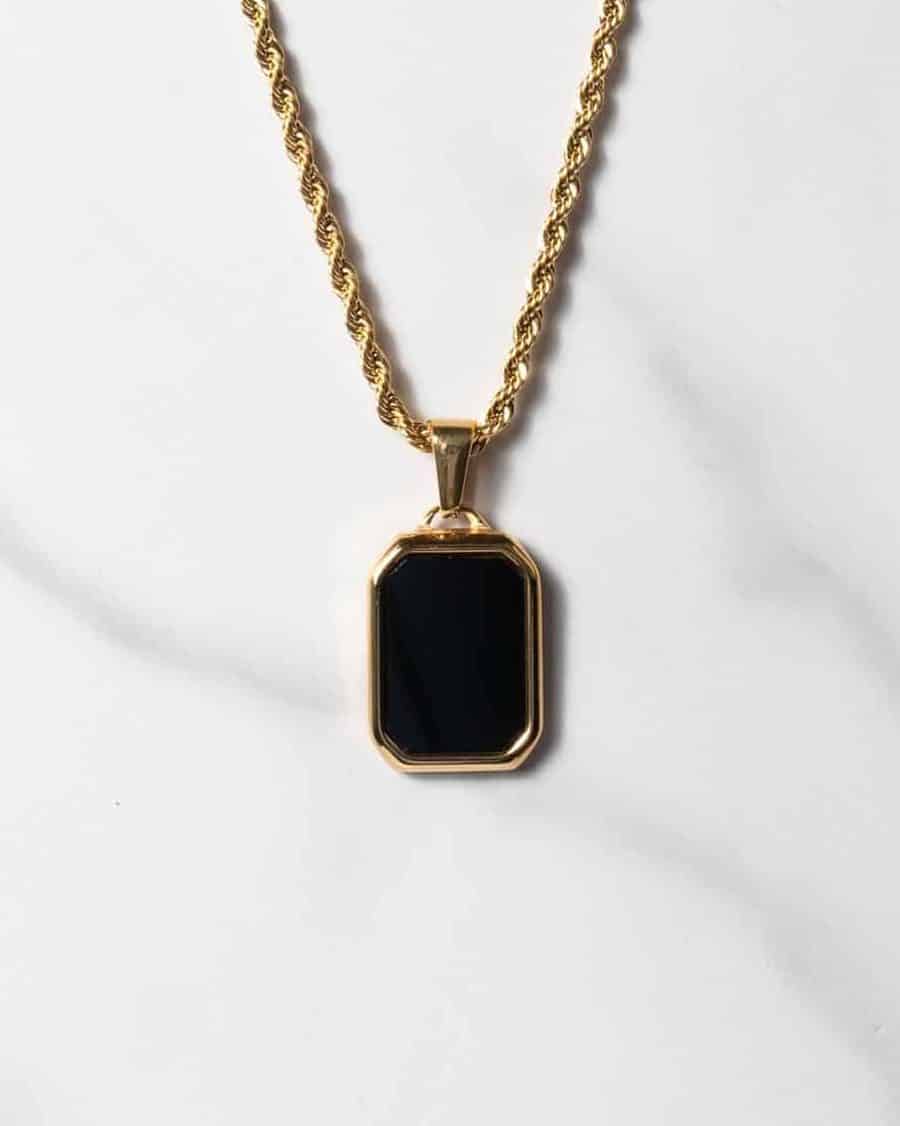 Ett halsband med repkedja i guld och ett rektangulärt hänge i svart onyx med konturer i guld, på en vit marmoryta.