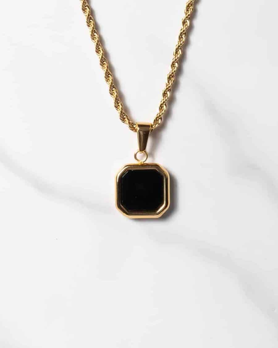 Halsband med repkedja i guld och ett åttkantigt hänge i svart onyx som omges av en guldram, elegant placerat på en vit marmorbakgrund.