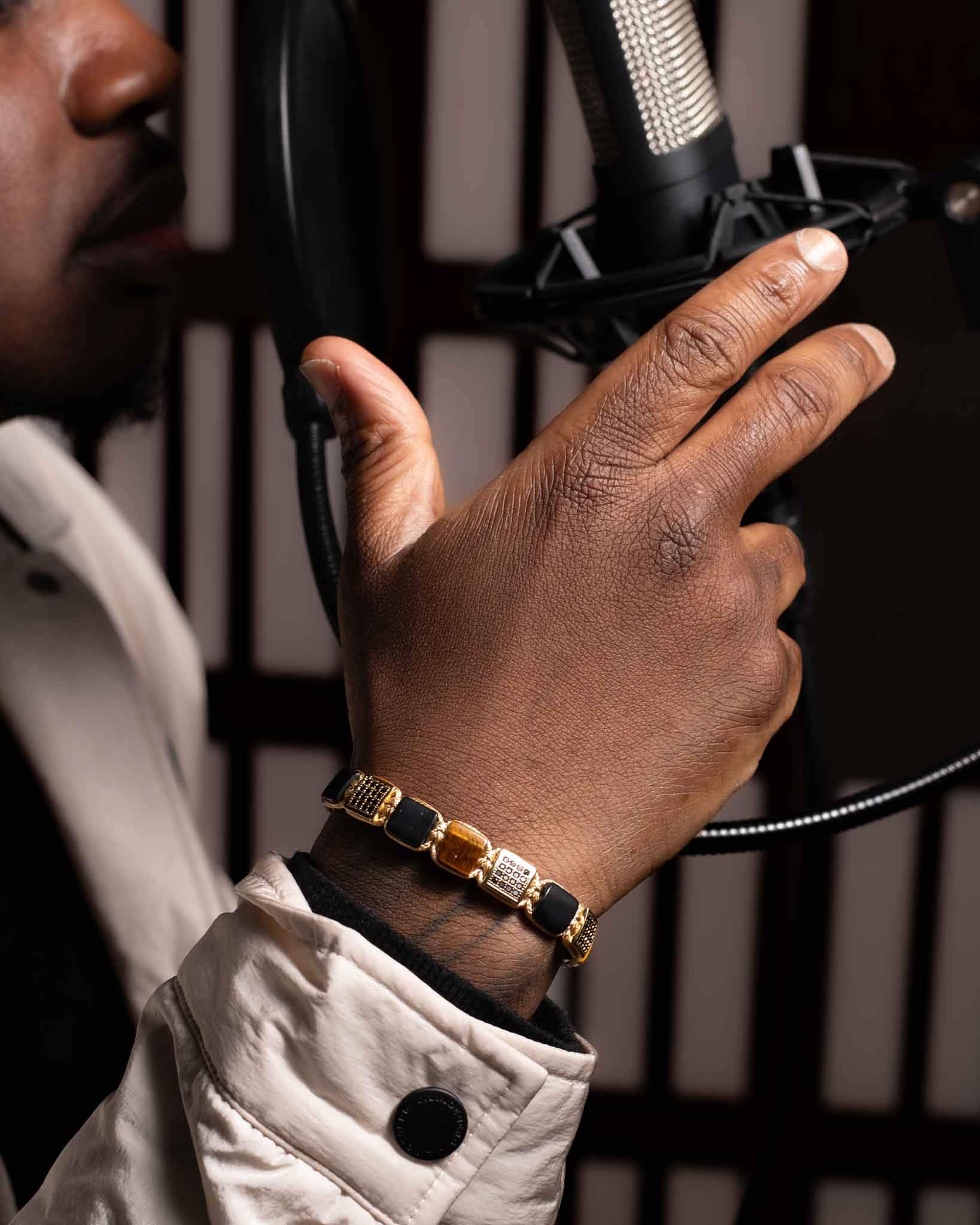 En person i beige jacka och Flatbead Marrakech-armband håller en mikrofon i en inspelningsstudio, med bara handen, en del av armen och undersidan av ansiktet synliga.