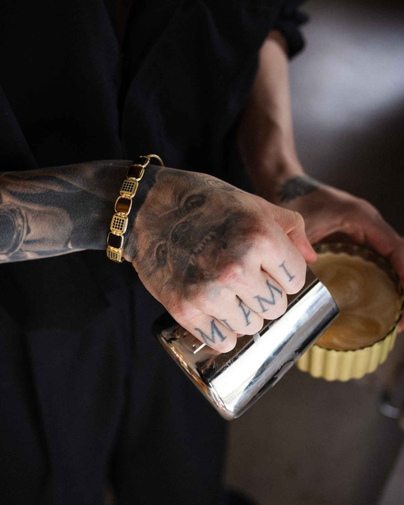 En person med en tatuerad hand som läser MIAMI och ett hundporträtt häller mjölk i kaffe, bär ett guldarmband och Flatbead Marrakech-armbandet.