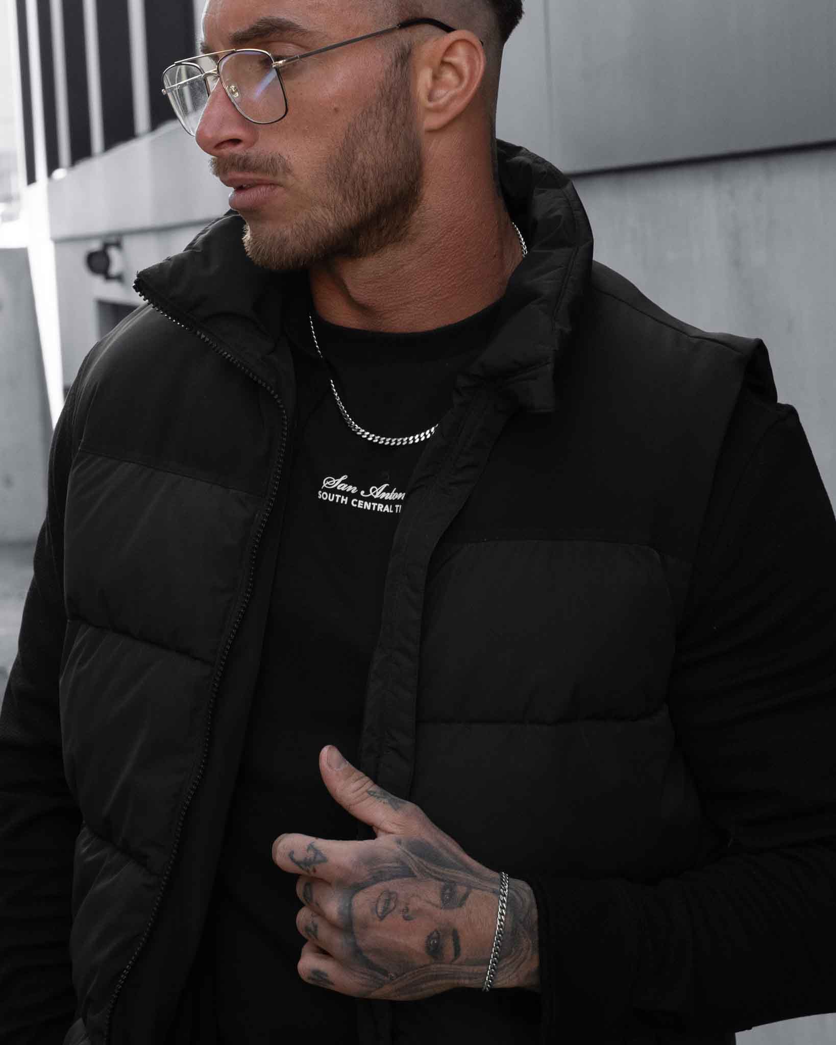 En man med kort hår, glasögon och handtatueringar bär en svart vadderad väst över en svart skjorta och ett silverhalsband på 5 mm med Pansarlänkinfattning medan han står utomhus vid en grå betongvägg.
