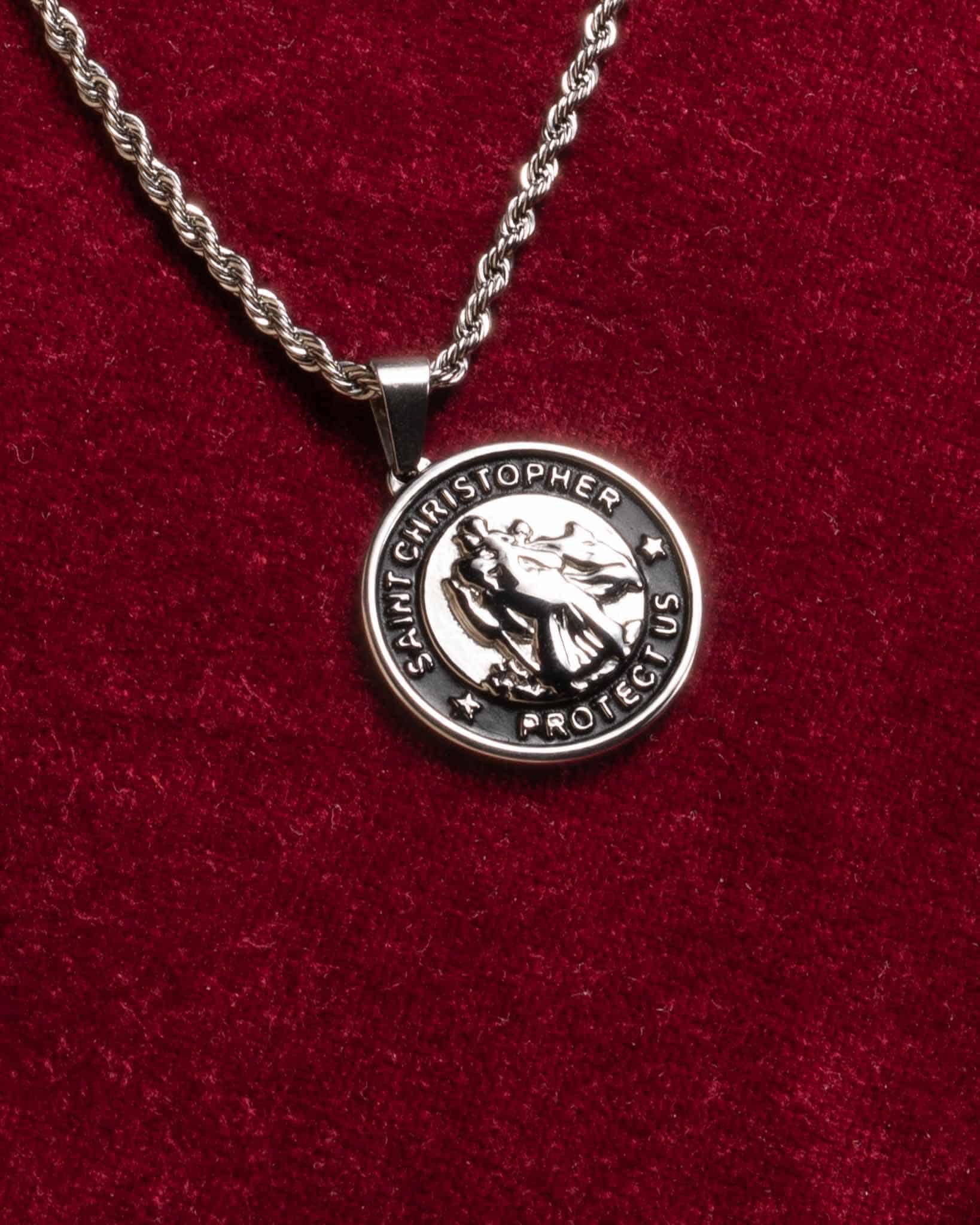 Ett Saint Christopher silverhalsband med hänge vilar på en texturerad röd tygbakgrund. Hänget har en graverad bild av Sankt Kristoffer och orden SAINT CHRISTOPHER PROTECT US runt kanten.