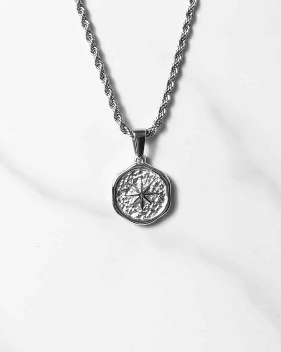 Ett Étoile Sauvage Silverhängehalsband med en texturerad rund medaljong med en kompassdesign, som visas på en tvinnad kedja mot en vit marmorbakgrund.