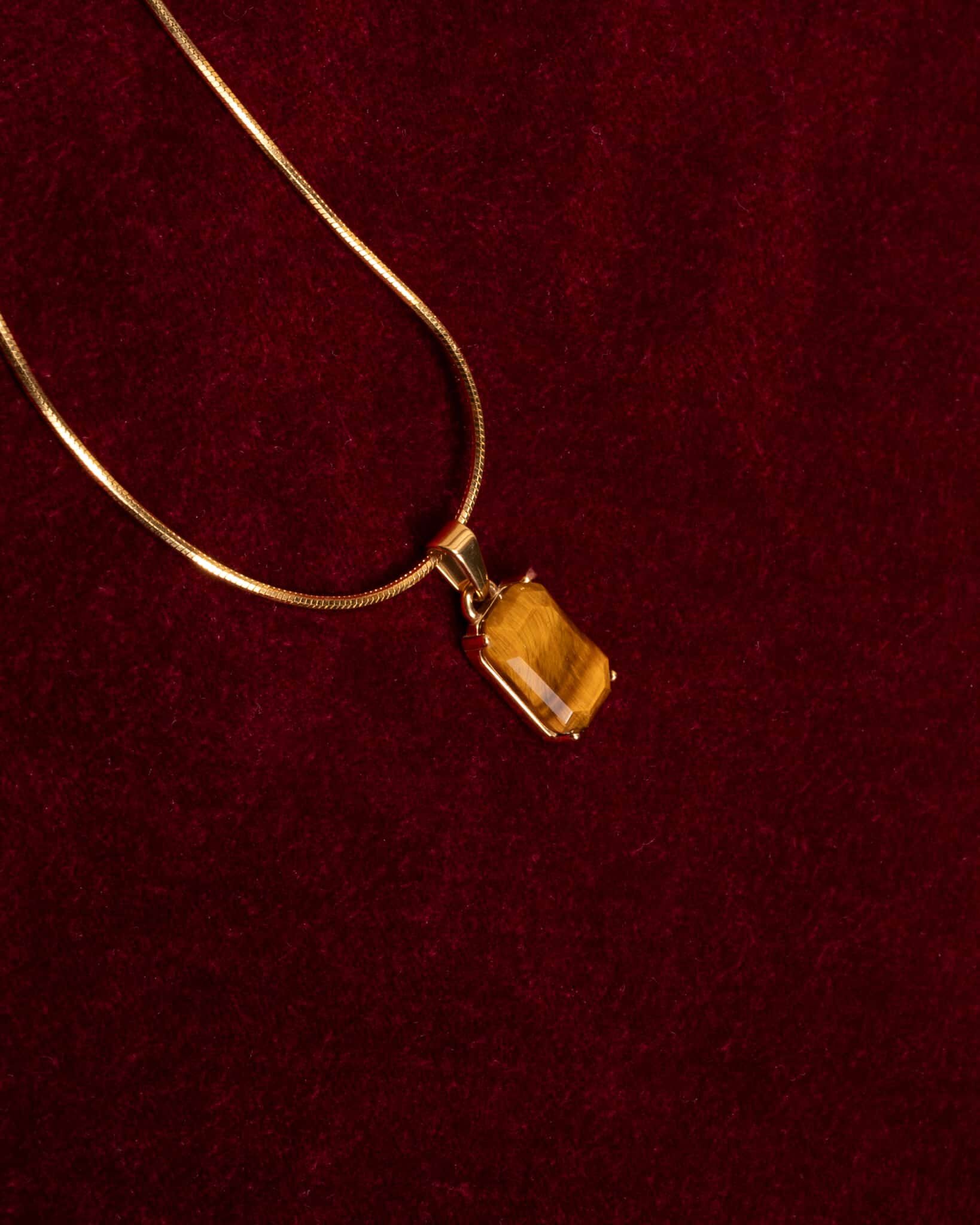 Ett guldhalsband med ett rektangulärt hängsmycke i form av en tigeröga visas på en texturerad mörkröd tygbakgrund.