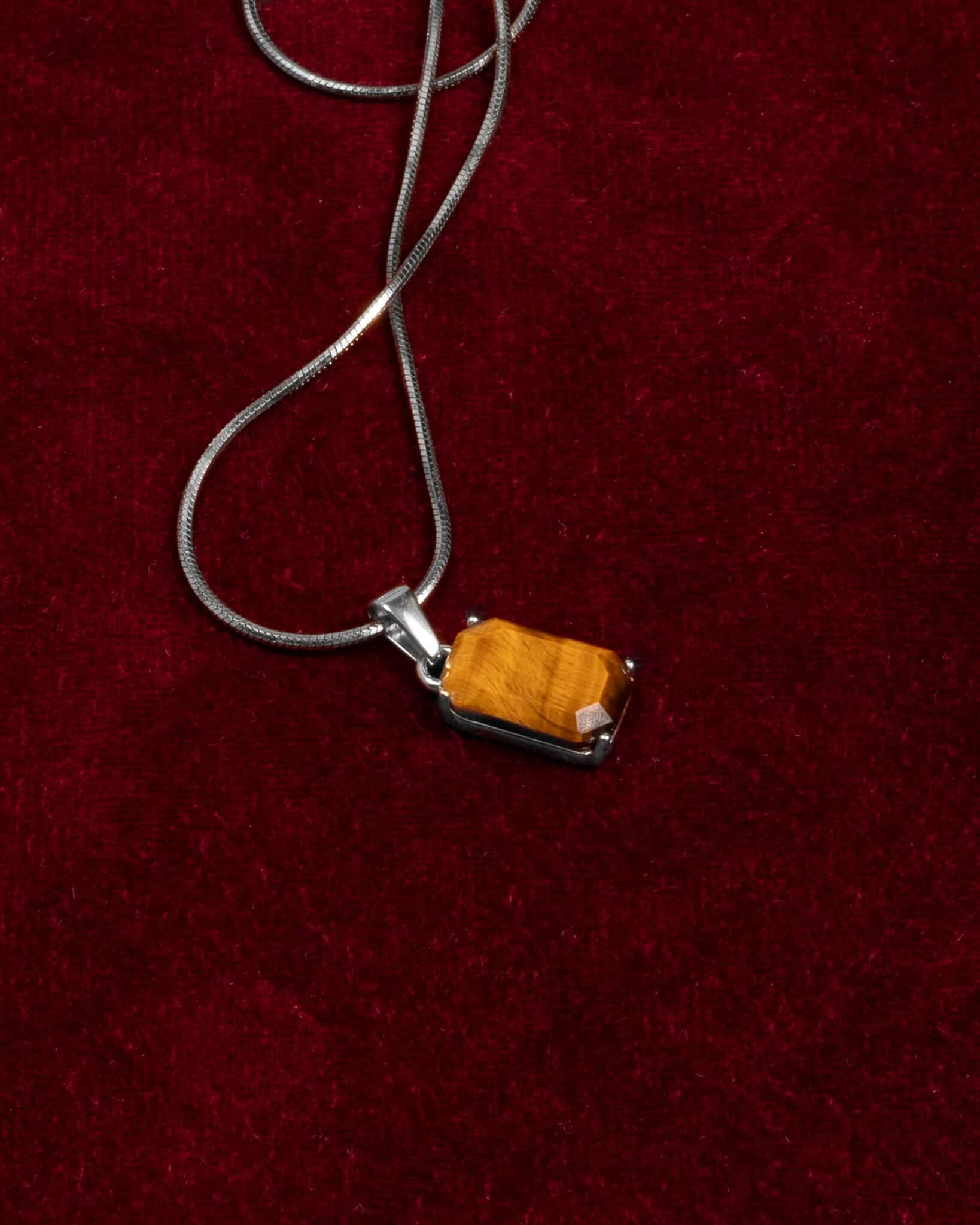 Ett silverhalsband med ett rektangulärt hängsmycke i form av ett tigeröga och en liten klar sten vilar på en riklig bakgrund av rött sammetstyg.