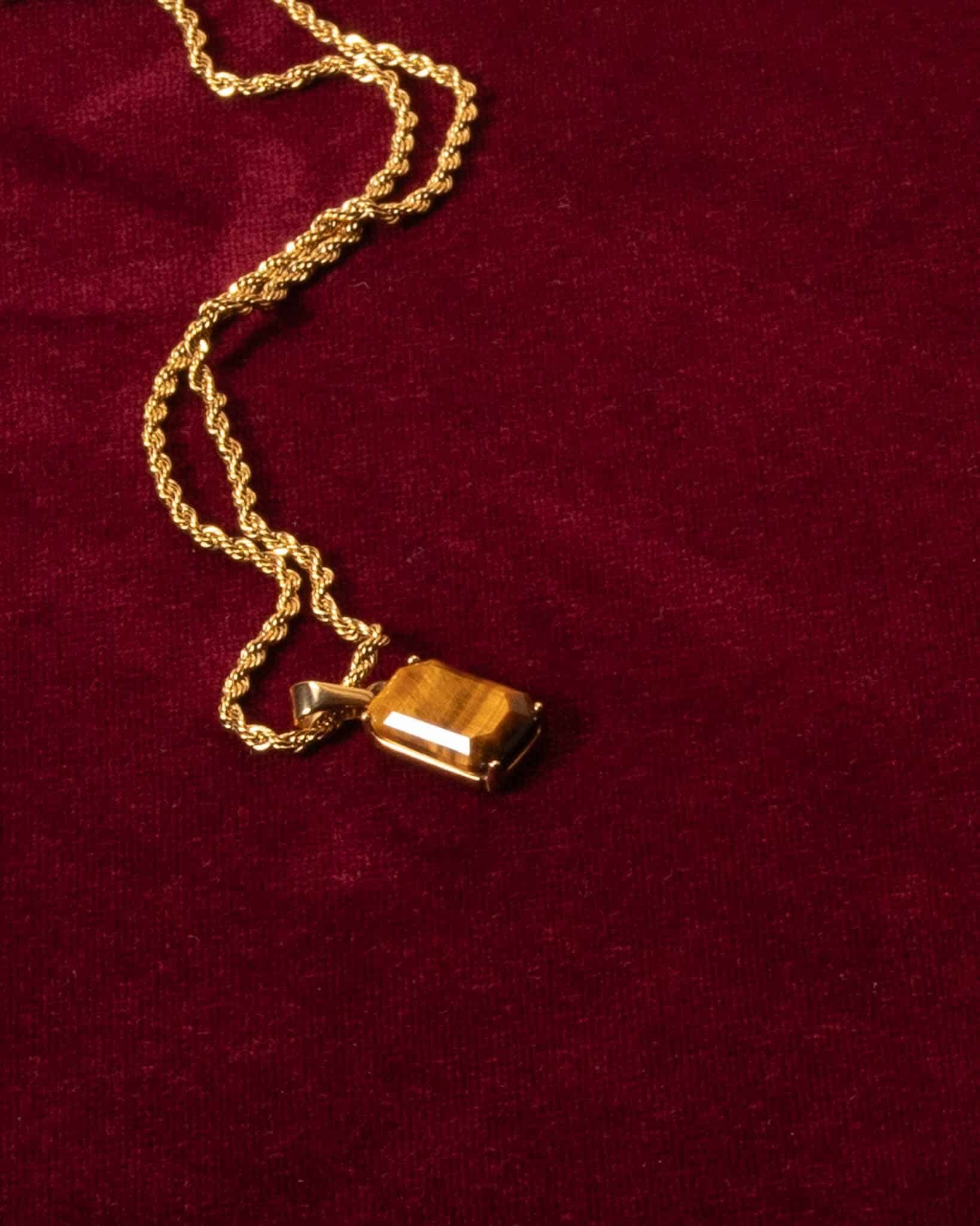 Ett Tiger Eye guldhalsband med ett rektangulärt brunt ädelstenshänge visas på vinrött sammetstyg.