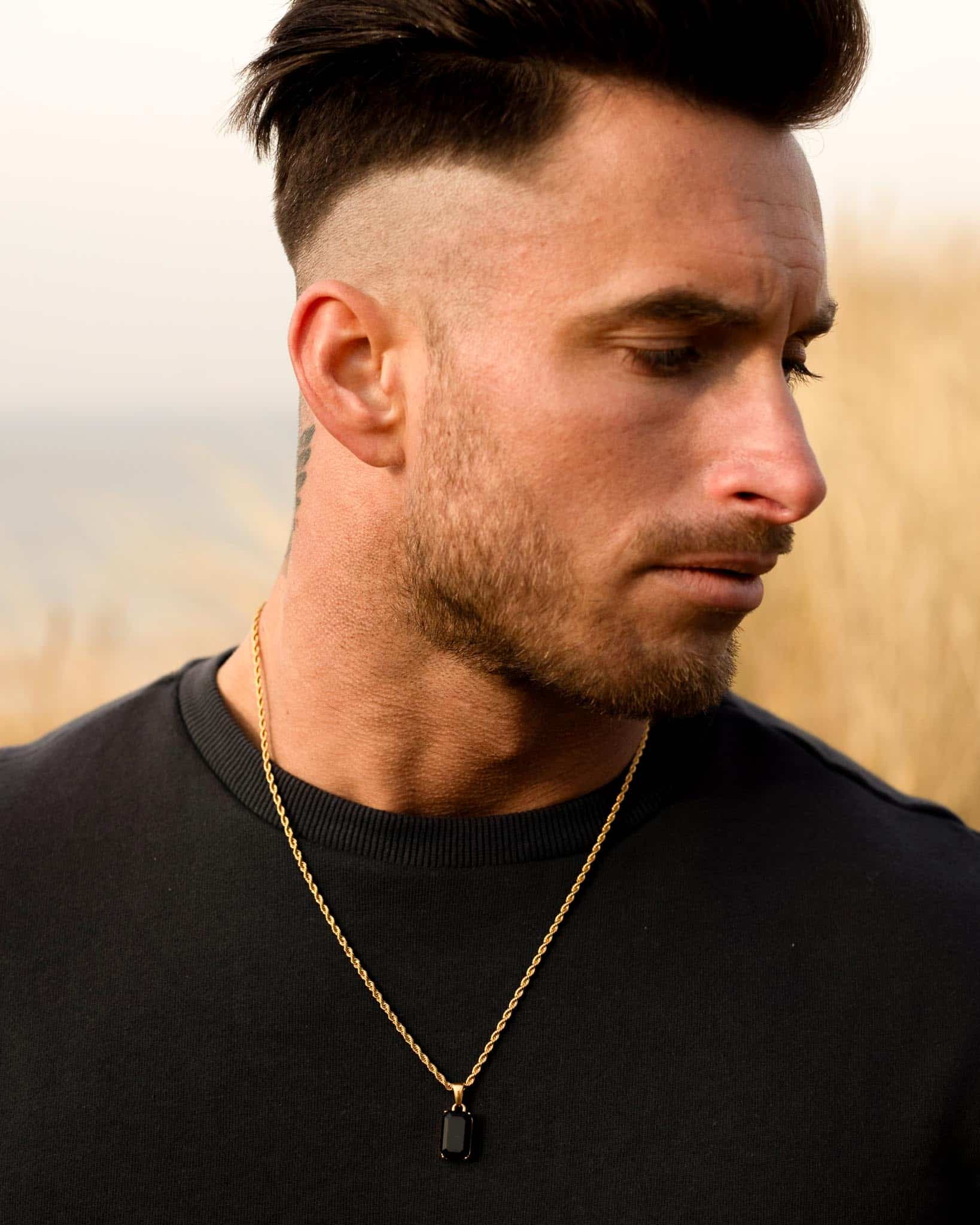 En man med kort mörkt hår och ett prydligt trimmat skägg tittar åt sidan, iklädd en svart skjorta och halsbandet Gemstone Black Onyx Guld med ett rektangulärt hänge, stående utomhus mot en suddig gräsbevuxen bakgrund.