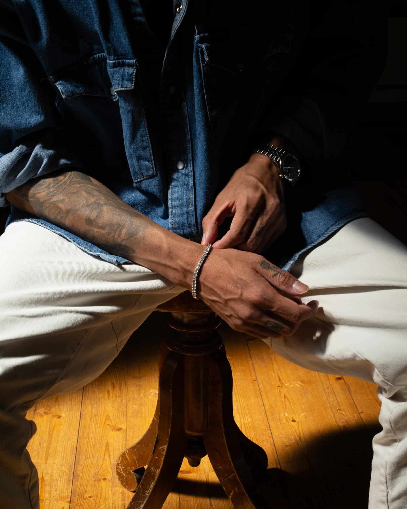 En person i blå jeansskjorta och vita byxor sitter på en träpall med händerna vilande i knät och visar upp ett tennisarmband, ett armbandsur och synliga tatueringar på armarna. Varmt ljus framhäver händerna och benen.
